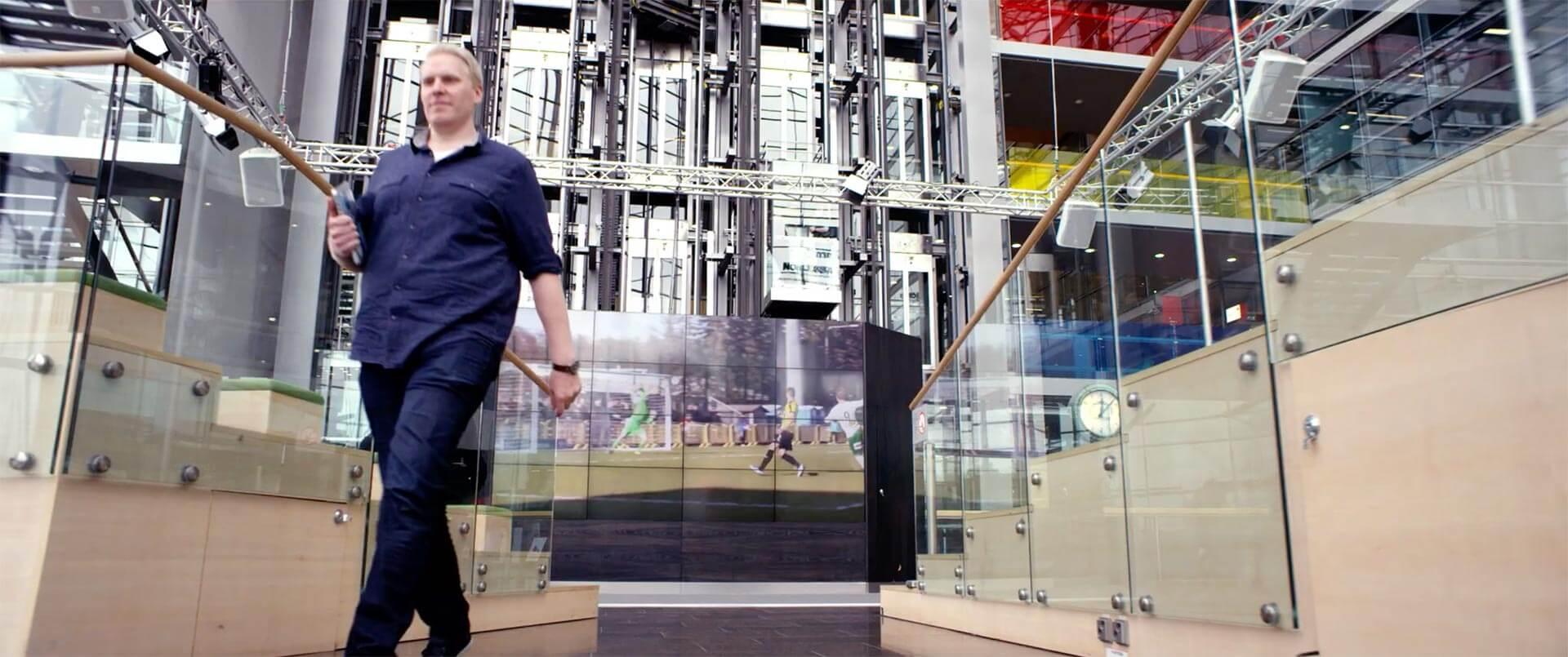  Man walking in office building with football streaming on big screen.