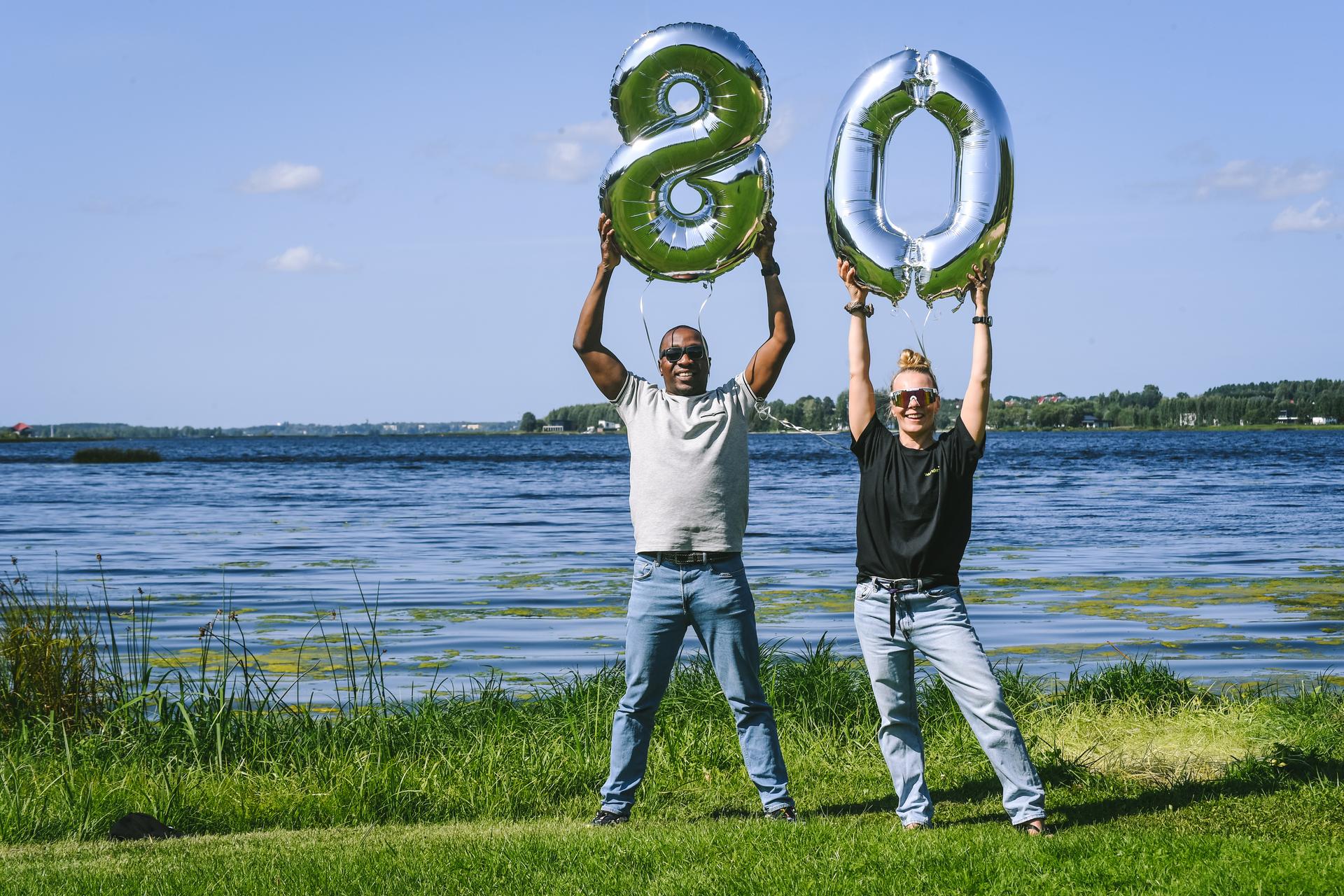 Two happy Mearra people standing next to the river on a sunny day, one of the persons holding foil balloon with number eight, another person holding number zero